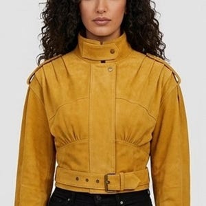 May include: A mustard yellow cropped jacket featuring a high collar with a snap button closure, shoulder epaulets, long sleeves, and a waist belt with a metal buckle. The jacket has a structured bodice with gathered detailing at the waist.