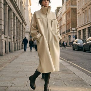 May include: A cream-colored long coat with a high collar and a full-length front zipper. The coat features two chest pockets with vertical zippers, long sleeves with buttoned cuffs, and a loose, straight-cut silhouette reaching mid-calf.