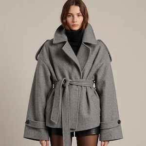 May include: A grey wool-blend jacket featuring a wide collar, long sleeves with buttoned cuffs, and a matching tie belt at the waist. The jacket has structured shoulders with epaulets and is worn over a black turtleneck and black shorts.