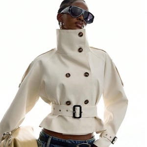 May include: A cream-colored cropped jacket featuring a high collar, double-breasted front with dark brown buttons, and a matching waist belt with a black buckle. The jacket includes shoulder epaulets and adjustable straps at the sleeve cuffs.