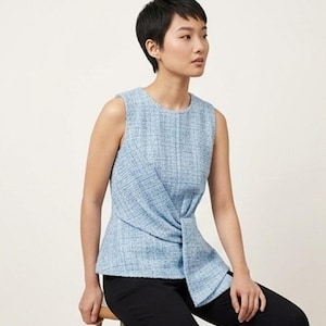 May include: A sleeveless light blue tweed top featuring a round neckline and a distinctive knotted detail at the front waist. The fabric has a textured, woven appearance with subtle white and darker blue threads throughout the material.
