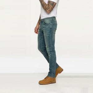 May include: A pair of slim-fit blue denim jeans with a faded wash, featuring a classic five-pocket design, a button fly, and metal rivets. The jeans are paired with a plain white short-sleeved t-shirt and tan leather lace-up ankle boots.