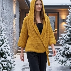 Puede incluir: Un cárdigan de punto color amarillo mostaza con capucha, mangas tres cuartos y un frente estilo cruzado que se asegura con un lazo a juego en la cintura. La prenda tiene un patrón de punto grueso y texturizado sobre una blusa y pantalones negros.