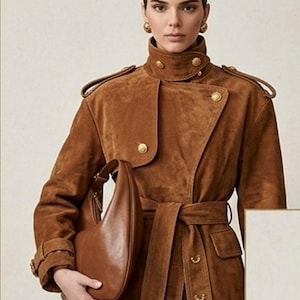 May include: A brown suede trench coat featuring a high collar, shoulder epaulets, and gold-toned buttons. The coat includes a matching waist belt and is paired with a smooth, crescent-shaped brown leather shoulder bag and round gold earrings.