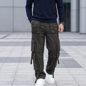 May include: Dark charcoal grey cargo trousers featuring a textured fabric, two front hip pockets with flaps, two large side cargo pockets with flaps, and additional lower leg pockets with hanging fabric straps, paired with a navy blue long-sleeve shirt.