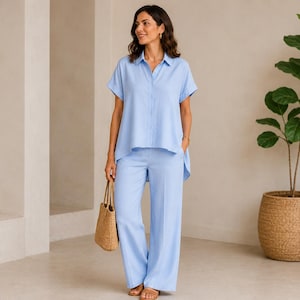 Può includere: Una camicia a maniche corte azzurra con bottoni, colletto e orlo asimmetrico, abbinata a pantaloni larghi azzurri coordinati. L'outfit è completato da una borsa in paglia color cuoio e sandali marroni aperti.
