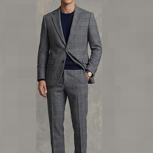 Pode incluir: Um fato cinzento aos quadrados composto por um casaco e calças a condizer com um padrão de xadrez cinzento escuro e preto. O fato é usado sobre uma camisola azul-marinho de gola redonda. Um relógio dourado com bracelete castanha é visível.