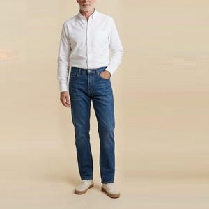 May include: A long-sleeved white button-down shirt with a collar and a single chest pocket, paired with medium-wash blue denim jeans and light-coloured trainers. The shirt is tucked into the jeans, which feature a classic five-pocket design.