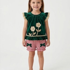 May include: A dark green velvet top with ruffled sleeves, gold lace trim, and two embroidered gold flower appliqués. The top is paired with red and white gingham shorts featuring a ruffled hem and two small dark green bows on the front.