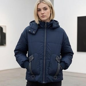 May include: A navy blue puffer jacket with a textured grid pattern, a full-length front zipper, and a hood. The jacket features two front pockets with dark grey trim, ribbed cuffs, and two metal D-rings attached near the bottom hemline.