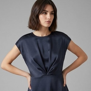 Op de afbeelding: Een donkerblauwe mouwloze jurk met een ronde halslijn en een geplooid, gedraaid knoopdetail in het midden van de taille. Het kledingstuk is gemaakt van een gladde, satijnachtige stof met een subtiele glans, wat zorgt voor een gestructureerd silhouet.