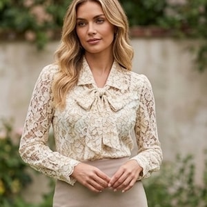 May include: A long-sleeved cream-coloured lace blouse featuring a pointed collar and a matching fabric bow tie at the neckline. The intricate floral lace pattern covers the entire bodice and sleeves, which are finished with buttoned cuffs.
