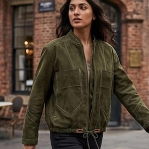 Puede incluir: Una chaqueta bomber de ante verde oliva con cuello alzado, cremallera frontal completa y dos grandes bolsillos de parche en el pecho. La chaqueta incluye detalles de costuras visibles y un dobladillo con cordón y cierres de cuero.