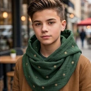 May include: A dark green infinity scarf featuring a repeating pattern of small, embroidered gold star-like symbols. The scarf is draped loosely around the neck, layered over a dark green shirt and a tan jacket with a visible front zipper.