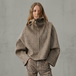 May include: A taupe-colored, textured zip-up jacket with a high collar and wide sleeves, paired with matching taupe pants featuring a subtle, dark, abstract pattern. The jacket has a central zipper closure and a loose, boxy fit throughout.