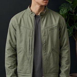 May include: An olive green utility jacket featuring a stand-up collar, a full-length front zipper, and structured paneling. The jacket includes two vertical chest pockets and two angled lower pockets with reinforced stitching details throughout.