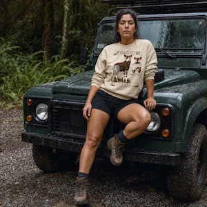 Puede incluir: Una sudadera beige de cuello redondo con una ilustración gráfica de un okapi y el texto "FIELD NOTES" y "OKAPI" impreso en la parte delantera. El diseño incluye dibujos detallados del animal en varias posturas sobre un fondo natural.