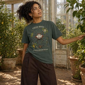 Op de afbeelding: Een donkergroen T-shirt met korte mouwen en een grafische print van een stekelige wielwebspin in een web, met de tekst "FIELD NOTES" bovenaan en "SPINY-BACKED ORB WEAVER" onderaan, gecombineerd met een donkerbruine broek.