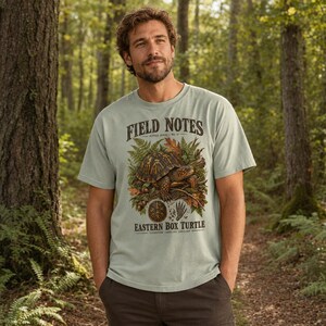 以下が含まれることがあります： セージグリーンの半袖Tシャツ。シダや葉に囲まれたトウブハコガメのグラフィックプリントが施されています。テキストには「FIELD NOTES」「EASTERN BOX TURTLE」「TERRAPENE CAROLINA CAROLINA」と記され、小さな亀の甲羅のアイコンが添えられています。