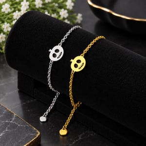 Könnte beinhalten: Zwei Kettenarmbänder, eines in Silber- und eines in Goldoptik, jeweils mit einem flachen, kreisförmigen Anhänger in Form eines stilisierten Totenkopfes mit zwei großen Augenhöhlen und einer genähten Mundpartie, versehen mit einem runden Endstück.