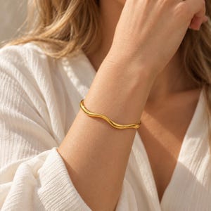 Può includere: Un bracciale rigido in metallo dorato lucido caratterizzato da un design a onda continuo, liscio e sinuoso. Il metallo presenta una finitura riflettente ad alta lucentezza che cattura la luce, creando una forma circolare semplice.