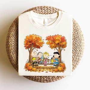 May include: A cream-colored t-shirt featuring a graphic print of the Peanuts characters sitting on a wooden bench under two autumn trees. The scene includes orange pumpkins on the ground, falling leaves, and the characters holding mugs.