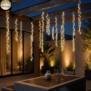 Op de afbeelding: Een reeks verticale lichtsnoeren met warmwitte ledlampen die aan een houten pergola hangen. De lampjes vormen meerdere gloeiende strengen die het terras verlichten, met een donkere tafel en twee wijnglazen in de voorgrond.