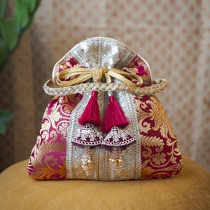 May include: A drawstring pouch bag featuring a deep magenta and gold floral brocade pattern. The bag includes a central silver panel with sequins, a braided gold handle, and two decorative dark red tassels with beaded accents hanging from the top.
