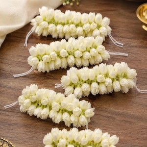 May include: Five individual garlands of white jasmine flower buds arranged in a staggered pattern. Each cylindrical floral strand is densely packed with small, cream-colored blossoms and features thin white thread ties extending from both ends.