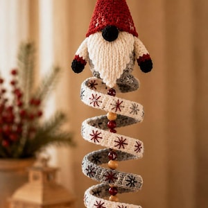 May include: A hanging crochet gnome ornament featuring a red pointed hat, a white beard, and a long, spiraling gray body decorated with embroidered dark red snowflake patterns. The spiral is accented with wooden beads and ends in a gray ball.