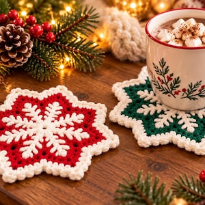 May include: Two star-shaped crochet coasters featuring a snowflake design. One coaster is red with a white border and snowflake center, while the other is dark green with a white border and snowflake center, supporting a white mug with red trim.