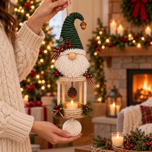 Puede incluir: Un adorno colgante de macramé con forma de gnomo que presenta un gorro texturizado de color verde oscuro con un pequeño dije de bellota, una barba de flecos color crema y una estructura de linterna central que sostiene una pequeña vela blanca. La pieza está decorada con ramas de pino, bayas rojas y pequeños lazos a cuadros, terminada con un gran pompón color crema en la parte inferior.