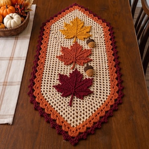 May include: A cream-colored crocheted table runner with a diamond shape, featuring three large maple leaf appliqués in yellow, orange, and dark red, along with two small brown crocheted acorns. The border is layered in orange and dark red yarn.