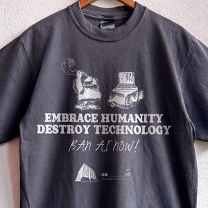 May include: A charcoal gray short-sleeve t-shirt featuring a white graphic of a caveman and a computer. The text reads EMBRACE HUMANITY DESTROY TECHNOLOGY, BAN AI NOW!, and includes a small building icon. The brand name SHAKAWEAR is printed below.