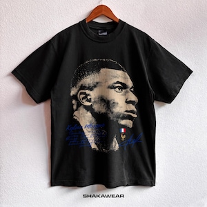 Può includere: Una t-shirt nera a maniche corte con un grande ritratto ad alto contrasto di Kylian Mbappé in tonalità beige e nero. Il design include testo blu, una piccola bandiera francese, un emblema con gallo dorato e una firma grafica blu.