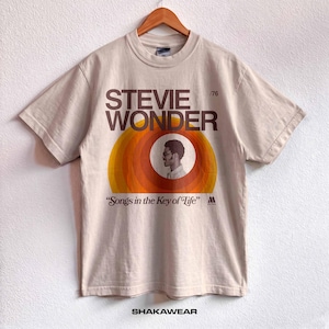 以下が含まれることがあります： ベージュの半袖Tシャツ。スティーヴィー・ワンダーのグラフィックと「STEVIE WONDER」、「/76」、「Songs in the Key of Life」の文字が、同心円状のオレンジと黄色の円の上に中央配置されています。下部にはブランド名「SHAKAWEAR」がプリントされています。