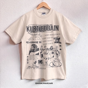 May include: A cream-coloured short-sleeve t-shirt featuring a black graphic print of Kurt Cobain. The design includes the text "KURT COBAIN," "NEVERMIND '91," "LOST INNOCENCE," "LOST YOUTH," "LOST MIND," "LOST VOICE," and "DON'T BE DISTRACTED" with illustrations.
