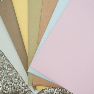 May include: A fan of different colored cardstock paper, including light pink, light blue, light brown, gold, and yellow, on a sparkly gold background.
