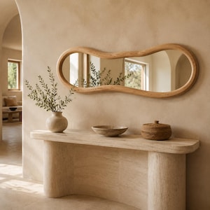 May include: A horizontal wall mirror with an irregular, organic, wavy wooden frame. The mirror reflects a room with a window and greenery. It is mounted above a curved stone console table holding a ceramic vase with branches, a shallow bowl, and a lidded basket.