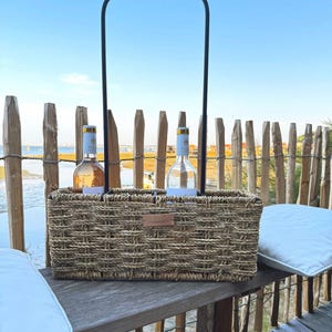 Peut inclure: Un panier rectangulaire en jonc de mer tressé doté d'une haute anse en métal noir, conçu pour contenir deux bouteilles de vin. Le panier présente une petite étiquette rectangulaire en cuir beige centrée sur le devant, avec une finition naturelle.