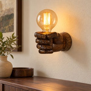 Puede incluir: Un aplique de pared que presenta una escultura de mano texturizada de color marrón oscuro que sostiene una bombilla grande, redonda y de cristal transparente con un filamento visible, emitiendo una luz cálida y suave.