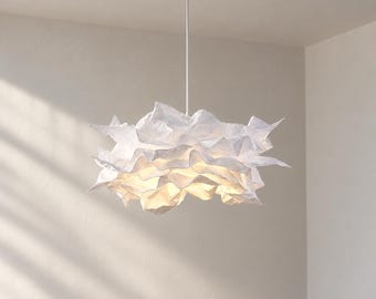 Może przedstawiać: Biała lampa wisząca z warstwowym, pofalowanym kloszem o fakturze przypominającej papier. Lampa jest zawieszona na cienkim białym pręcie przymocowanym do okrągłej podsufitki, emitując ciepłe światło wydobywające się z wnętrza fałd.
