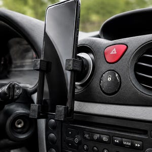 May include: A black smartphone mounted in a vertical car air vent holder. The holder features four square-shaped grips securing the device. The car dashboard includes a red hazard light button, a central lock button, and a radio control panel.