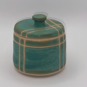 May include: A cylindrical ceramic jar with a matching lid featuring a knob handle. The exterior is finished in a teal glaze with a cream-colored grid pattern of intersecting lines that wrap around the body and the lid of the container.