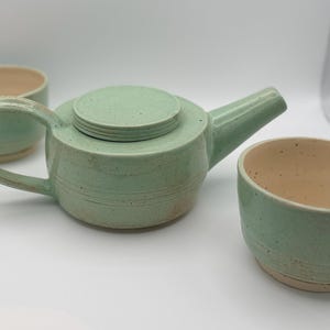 May include: A ceramic tea set consisting of a teapot with a lid and two matching cups. The pieces feature a speckled light mint green glaze on the exterior and a smooth, light beige interior. The teapot has a curved handle and a short spout.