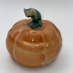 May include: A small, round, ceramic pumpkin figurine with a glossy orange finish and a textured surface. The pumpkin features a curved, dark green stem at the top, with visible ridges running down the sides of the body, creating a realistic shape.