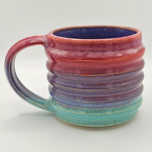 May include: A ceramic mug featuring a ribbed, cylindrical body with a gradient glaze that transitions from a vibrant red at the top to purple in the middle and a bright turquoise at the base. The interior is a deep purple hue.
