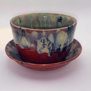 May include: A ceramic bowl and matching saucer set featuring a deep red base glaze with a decorative drip pattern in shades of cream, blue, and black. The glaze has a glossy finish, and the bowl has a wide, open top with a smooth, rounded rim.