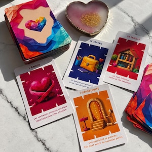 Op de afbeelding: Een set orakelkaarten met een geometrisch hartontwerp op de achterkant, vergezeld van vier losse kaarten met de titels L'AMOUR, LE TRAVAIL, LA FAMILLE en L'OPPORTUNITÉ, elk voorzien van kleurrijke illustraties en Franstalige beschrijvingen.