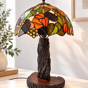 Op de afbeelding: Een tafellamp met een glas-in-loodkap met een kleurrijk patroon van oranje, groene en gele glazen stukjes. De lampvoet is ontworpen als een gestructureerde boomstam en rust op een platte, ronde houten schijf als voet.